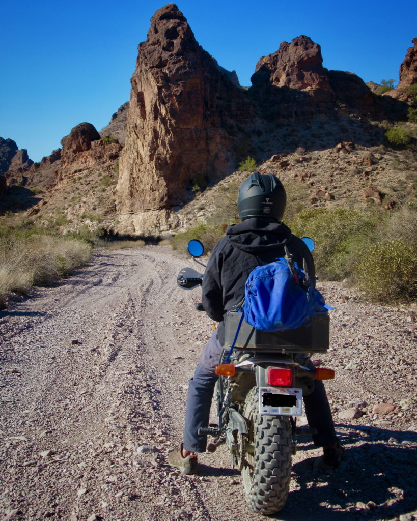 Man on motorbike facing away from camera with blue backpack on gravel road. Rugged brown peaks in background.
