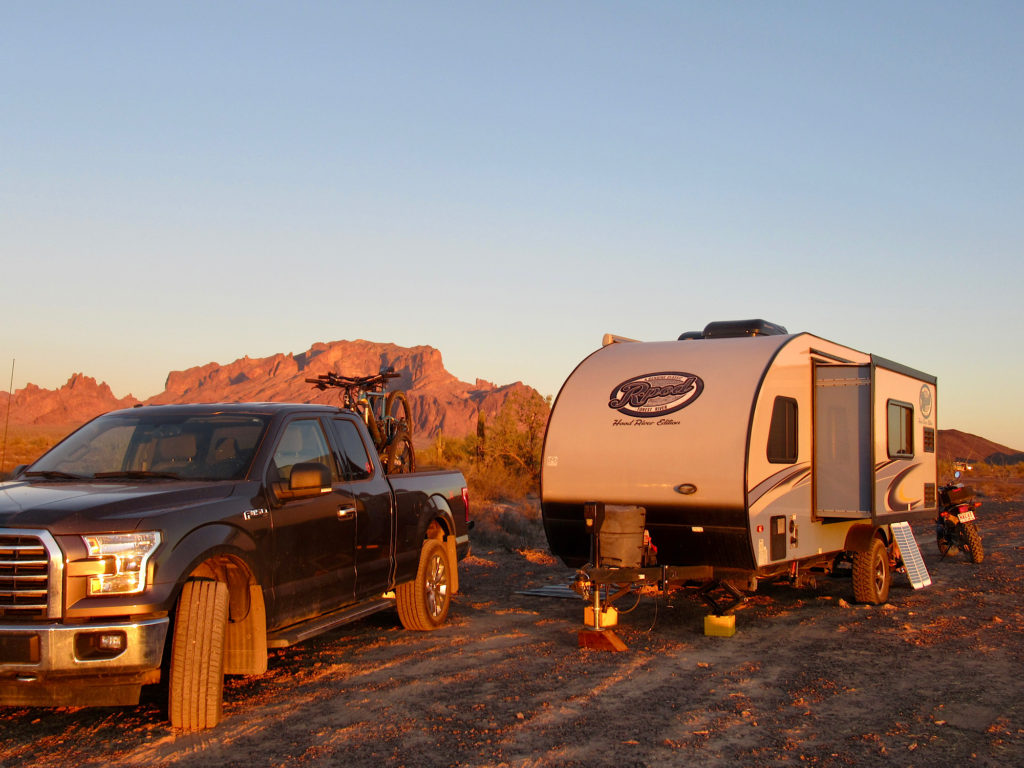 Grey pickup up and white and blue travel trailer in red glow of sunset with mountains in distance.