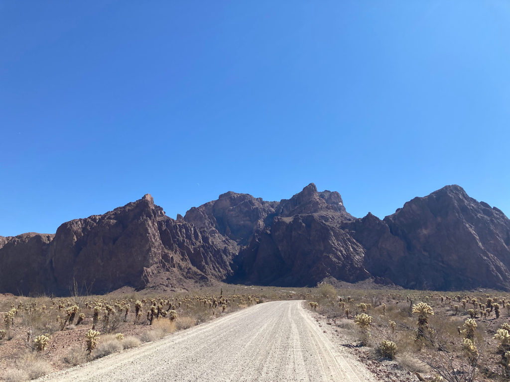 Wide gravel road leading directly to  dark brown jagged mountains.