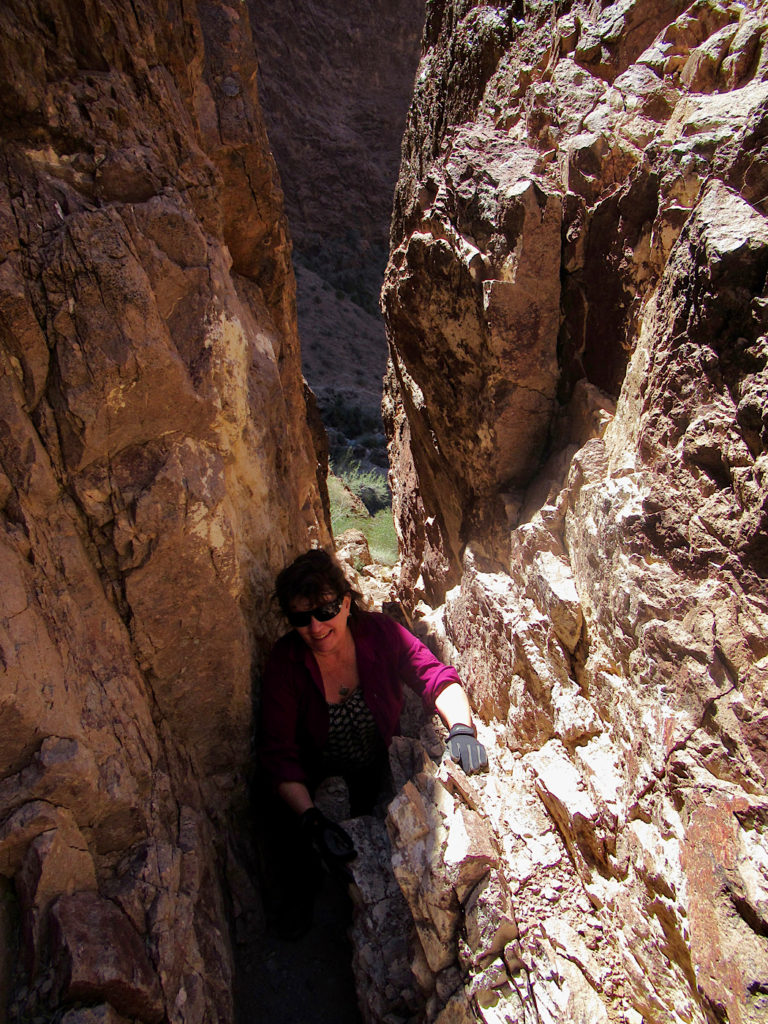 Woman in black ball cap and purple shirt climbing up through narrow slot in a canyon.