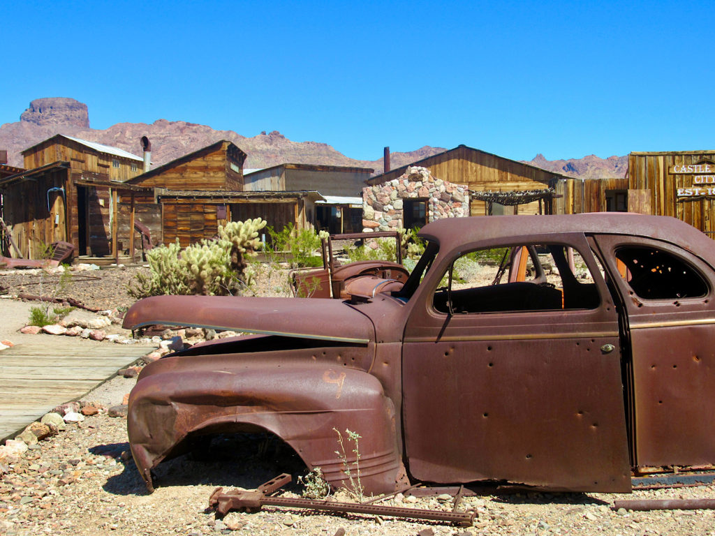 Rusting body of old car in front of wooden buildings.