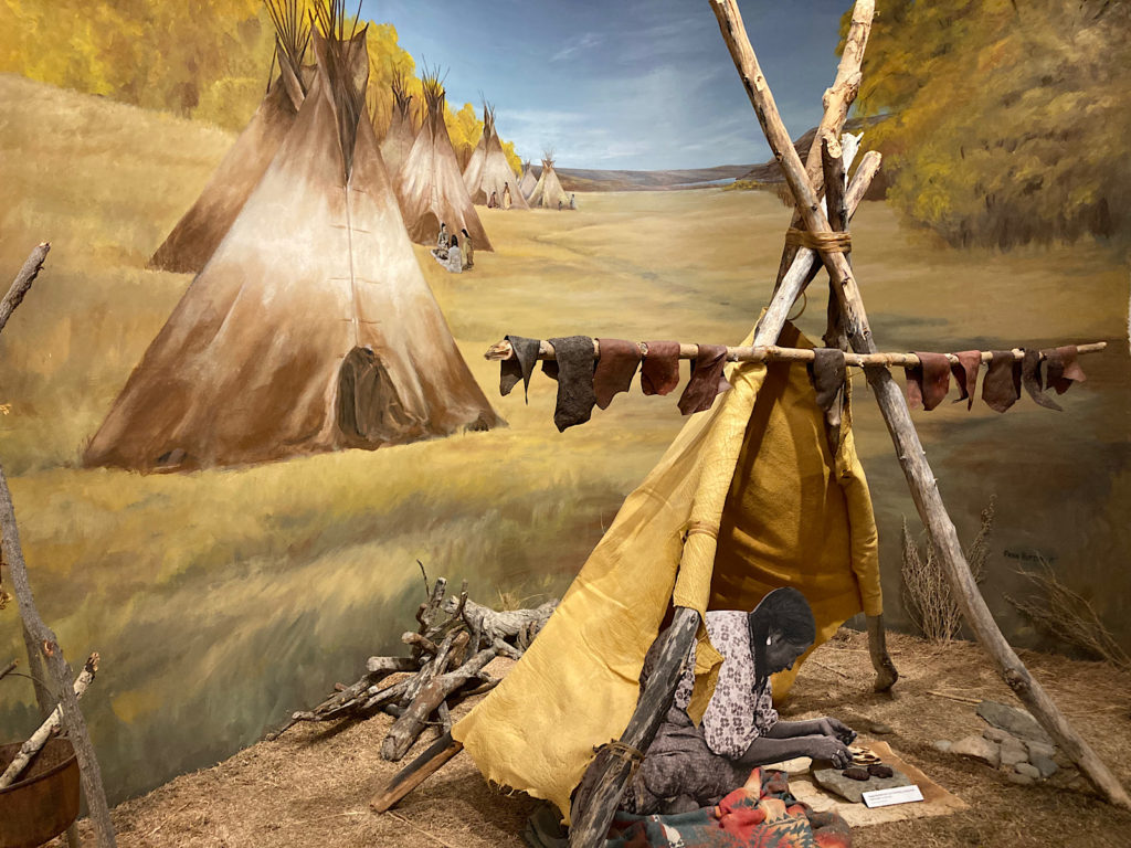 Museum display showing a painted wall with a row of teepees on the prairies and a physical teepee display in foreground.