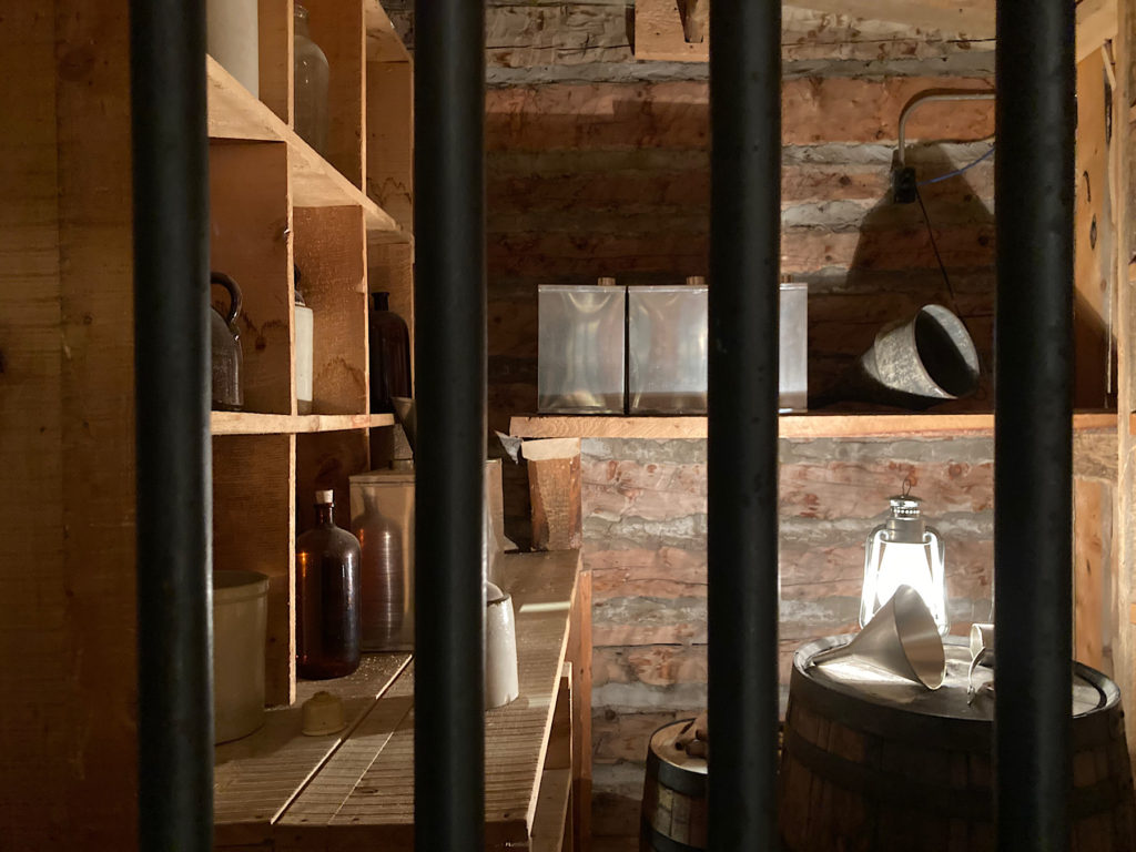 Metal jail cell bars protect the whisky bottles and tins on shelves with a lamp sitting on a barrel lighting up the wood walls.