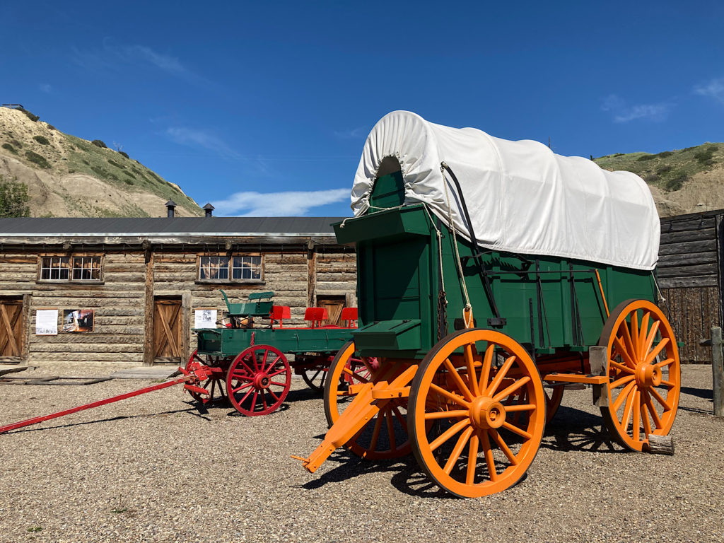 Two brightly painted green wagons, one with orange wheels and a white canvas roof and the other with no roof and red wheels in front of log building.