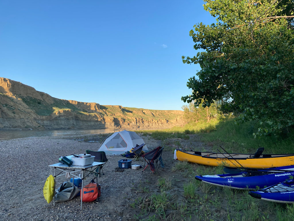 Yellow canoe and two blue inflatable kayak pulled up next to trees with metal table, folding chairs and tent set up on gravel beach next to river with brown cliffs on opposite side.