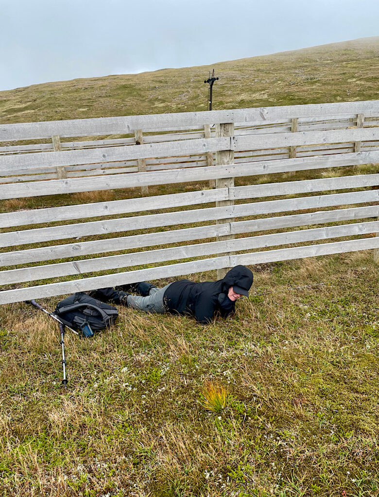 Woman in black raincoat and grey pants crawling under wooden fence on a day hike in Cairngorms National Park.