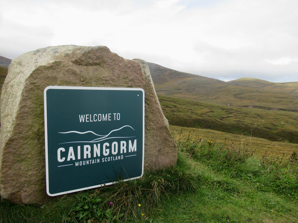 Welcome to Cairngorm Mountain Scotland written in white on dark green sign mounted on large rock with mountain moorlands in background.