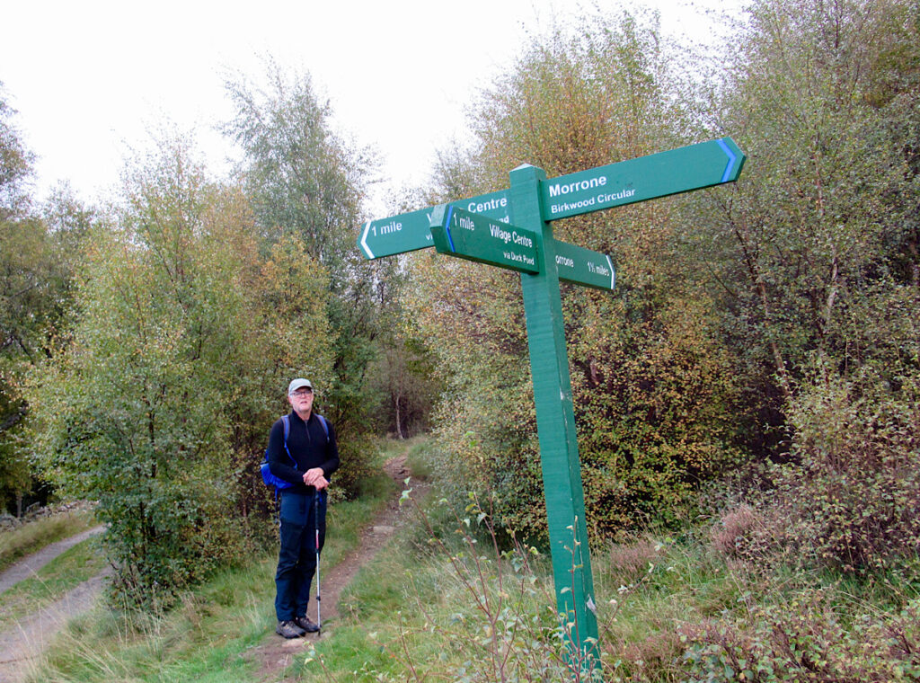 Man in dark shirt and pants standing with hiking pole looking at green sign pointing to Morrone Birkwood Circular trail.