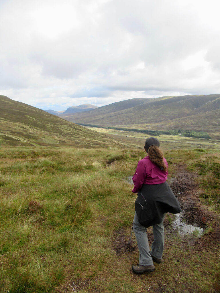 Woman in purple shirt, grey pants and black coat tied around waist looking out at rolling hills and moorland.