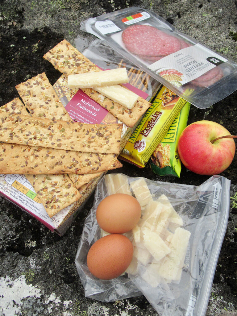 Eggs, cheese, apple, crackers, granola bars and salami laid out on a rock.