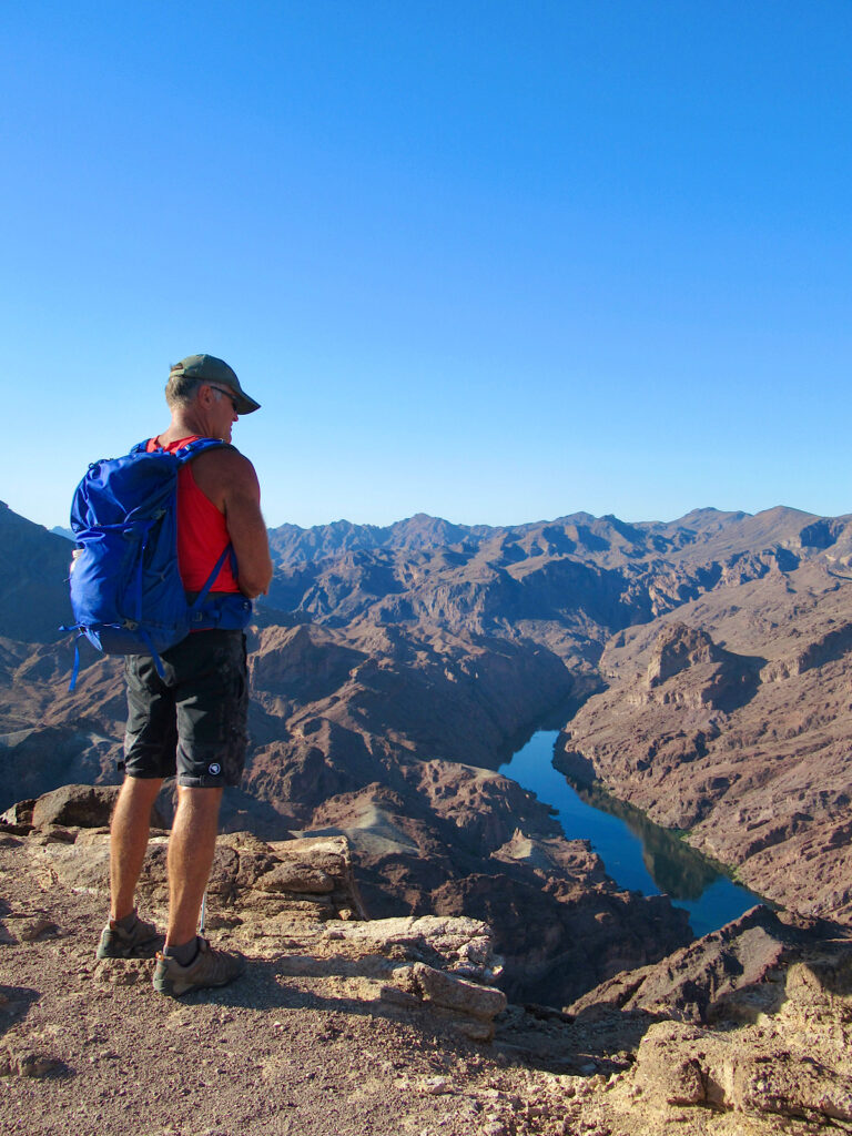 Male hiker in red shirt, grey shorts, ball cap and wearing blue backpack standing up on a red rock cliff above a river.