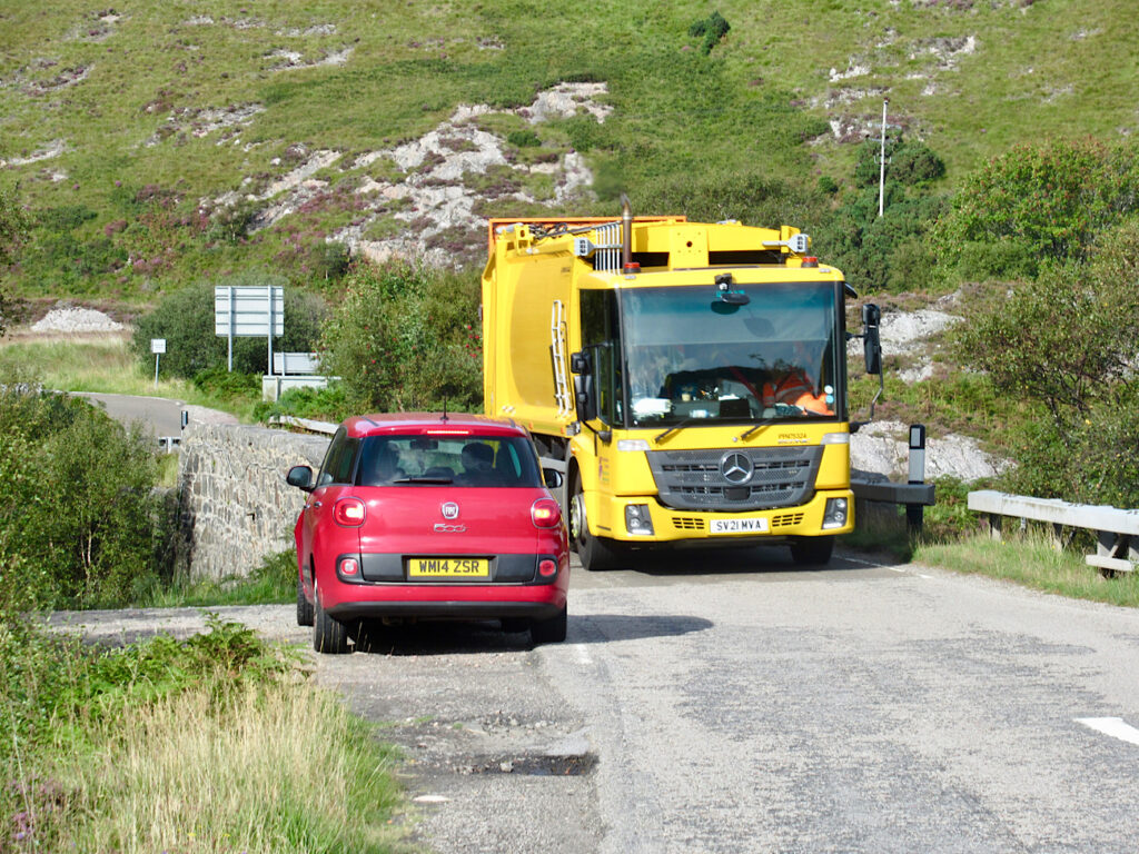 Red car pulled to side before narrow bridge filled with yellow truck.