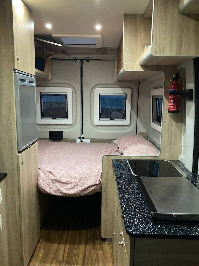 Interior of a camper van rental in Scotland with silver stovetop and fridge, black counters and light brown cupboards and pink comforter and pillows on rear bed.