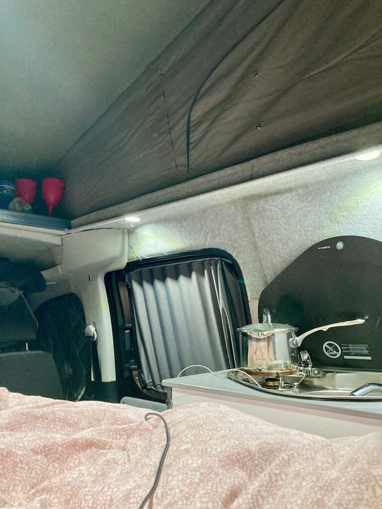 Inside a tiny camper van with a pot on a one burner stove next to sink and bed.