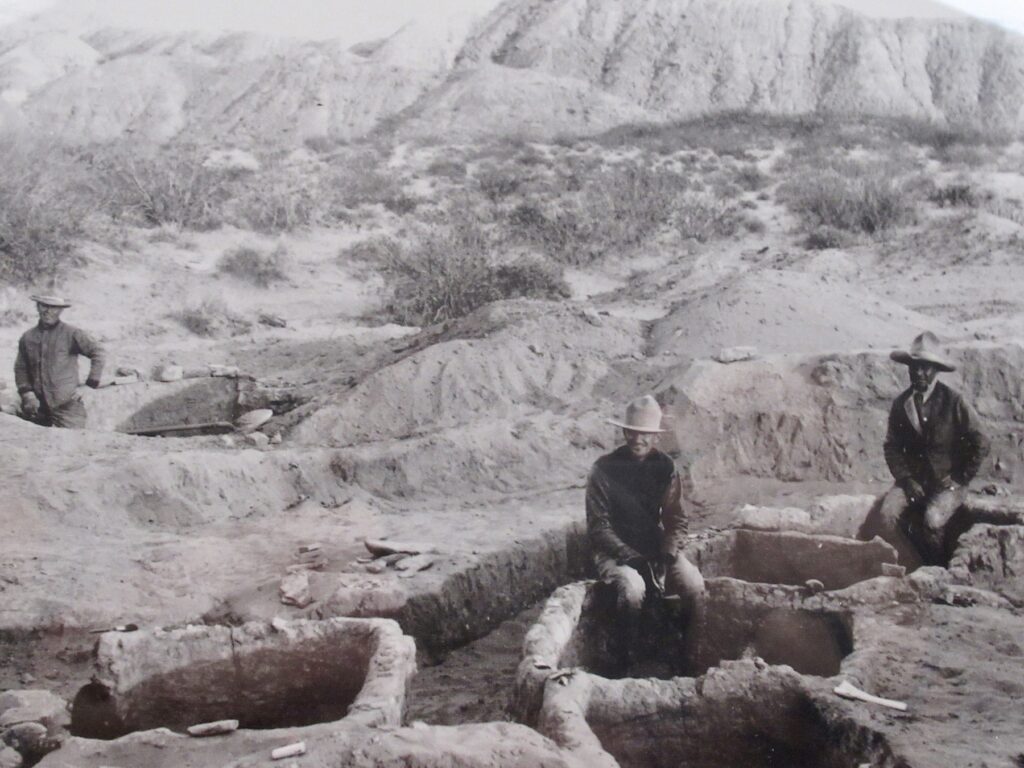 Black and white photo of three men wearing long-sleeved coats, long pants, and Stetson hats sitting on walls of archaeological dig in the desert with mountains in distance.