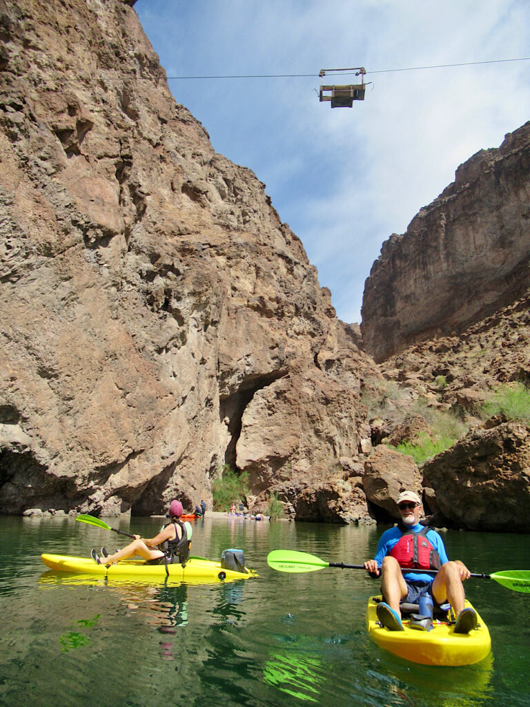 Man and woman on yellow sit on top kayaks holding green paddles on green river below steep brown canyon walls. Historic wooden tram cable car high above.