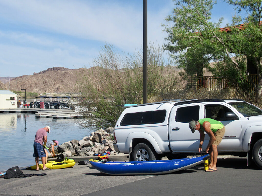 Man standing over blue inflatable kayak next to white pickup truck. Another couple with yellow sit on top kayaks beside water's edge.