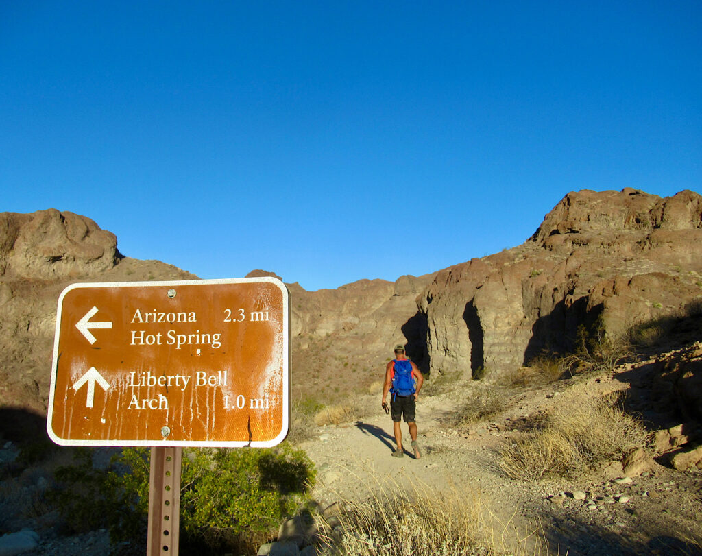 Man with blue backpack and black shorts hiking up trail beneath brown cliffs. Blue sky overhead. In lower left corner of picture is a brown trail sign with white lettering and arrows. It reads: Arizona Hot Spring 2.3 mi and Liberty Bell Arch 1.0 mi.