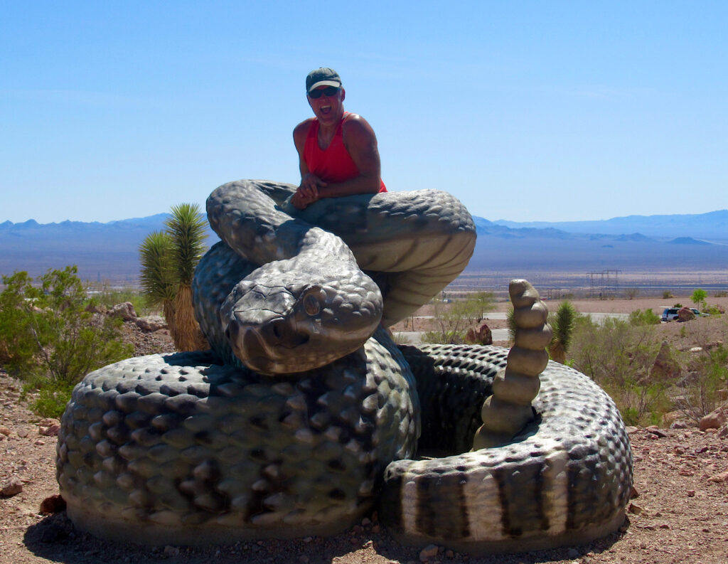 Man in red tank top wearing a green ball cap inside coils of massive rattlesnake sculpture.
