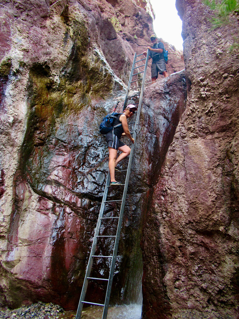 Woman in shorts descending metal ladder with man in shorts standing at top beside red rocks of canyon wall.