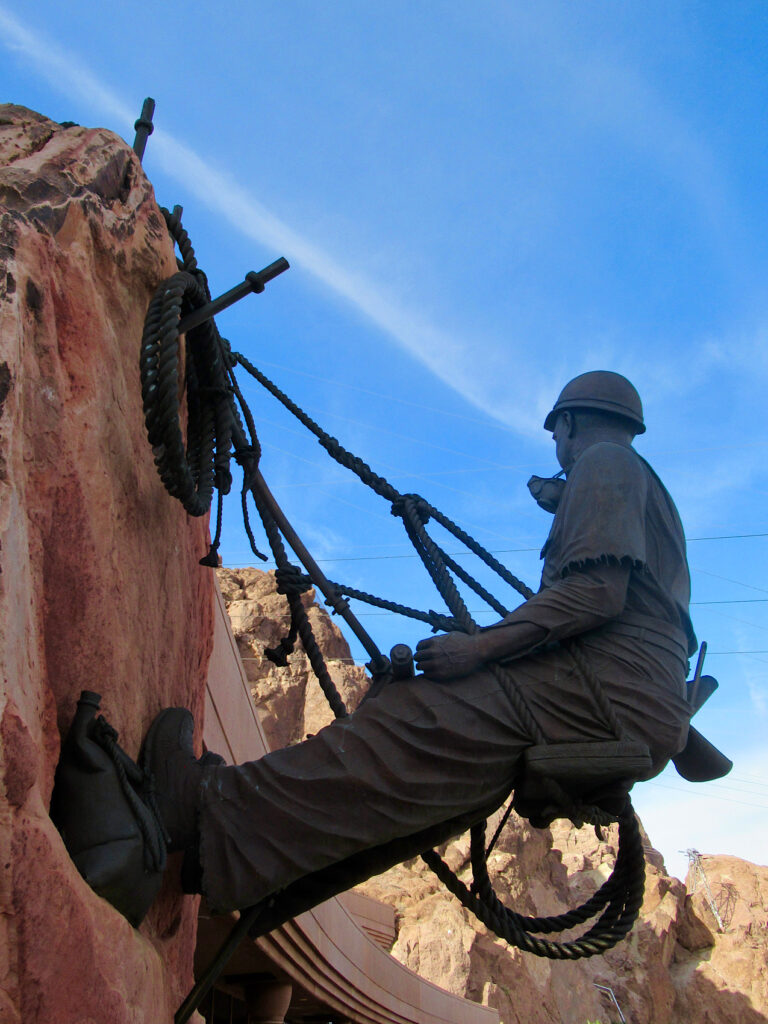 Bronze sculpture of a man held by ropes as he leans back from red rock wall.