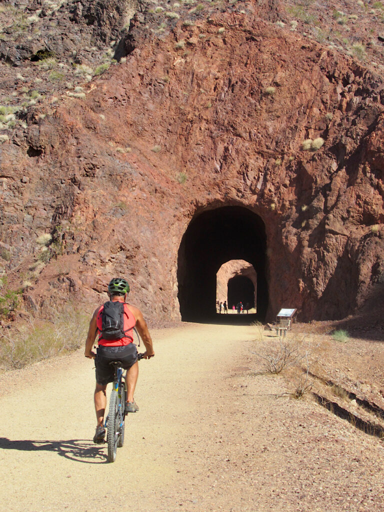 Man on bicycle riding towards a tunnel carved out of red rock.