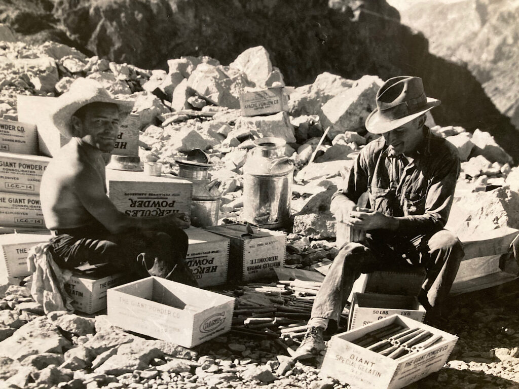 Black and white photo of two men wearing wide brim hats sitting on wood boxes labelled: The Giant Powder Company.