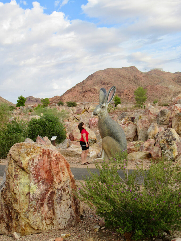 Woman in red shirt and black shorts standing in front of a sculpture of a jack rabbit that is twice her height. Red rocks in foreground and background.