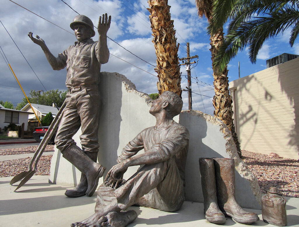 Bronze statues of two men, one leaning against a cement wall, wearing a hard hat and rubber boots with his hands up in the air. The second man sitting down, legs crossed looking up at first man. A pair of rubber boots and a canteen sit beside him. Palm trees in background.