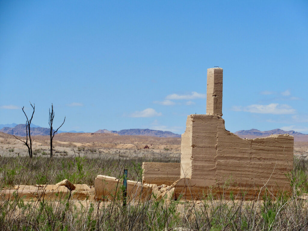Tall pale brown adobe chimney and wall with fallen sections in front and low brown cliffs and darker mountains in distance under blue sky.