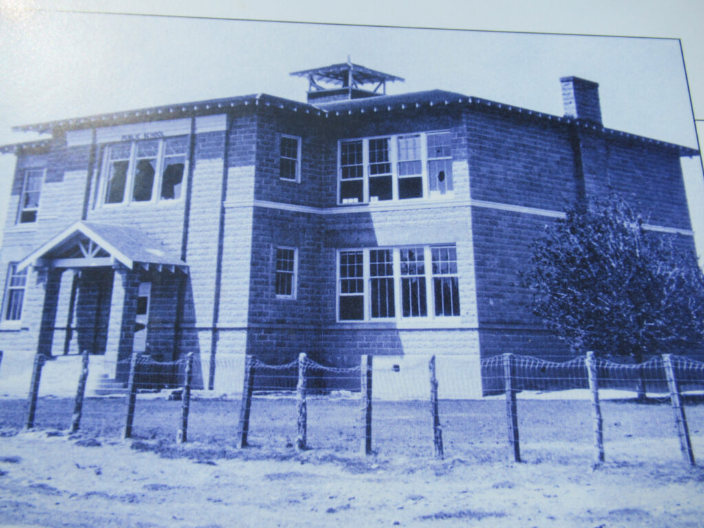 Black and white image of square, two-storey, brick building surround by page wire fence.