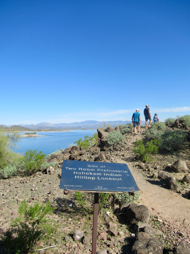 Two men and a woman in the distance cresting a hill above a lake with a sign in the foreground reading: "Site of Two Room Prehistoric Hohokam Indian Hilltop Lookout."