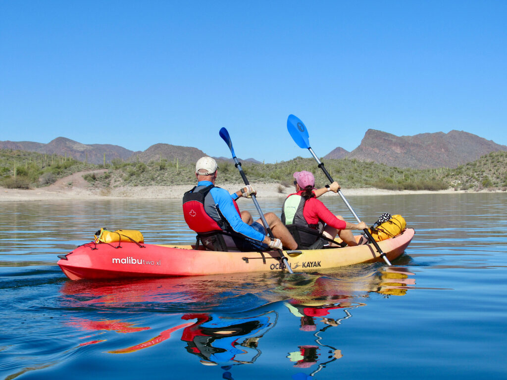 Man and woman in an orange and red sit on top kayak paddling on deep blue calm water with desert hills in background under blue sky.