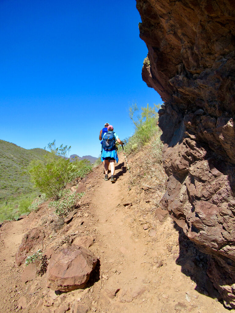 Man and woman hiking up steep, narrow brown dirt path below dark brown cliff under blue sky.