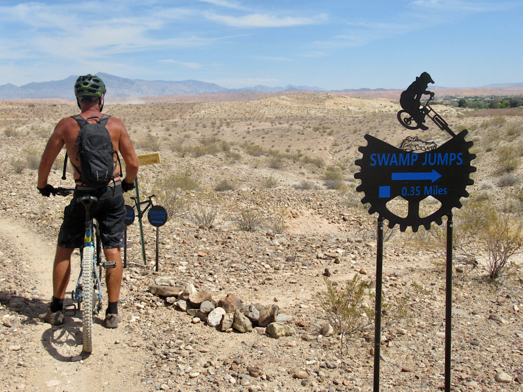 Man on mountain bike standing near trail barricaded with rocks. Sign reads" Swamp Jumps 0.35 miles" and show a cut out of a mountain biker on a jump.