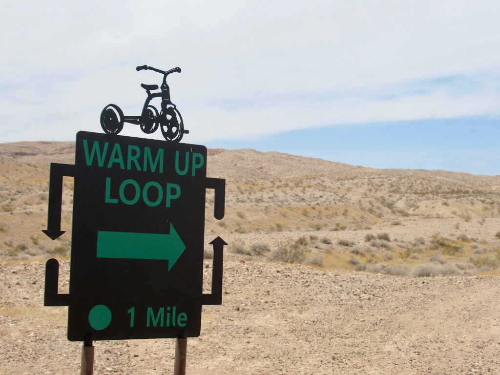 Metal sign in foreground with rocky desert in background. Sign reads: Warm Up Loop 1 mile. Metal cutout of a tricycle on top.