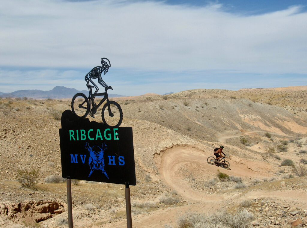 Metal sign with cutout of a skeleton wearing a helmet and riding a bicycle on top of metal sign titled "Ribcage". Man in background riding a bike down a winding hill trail.