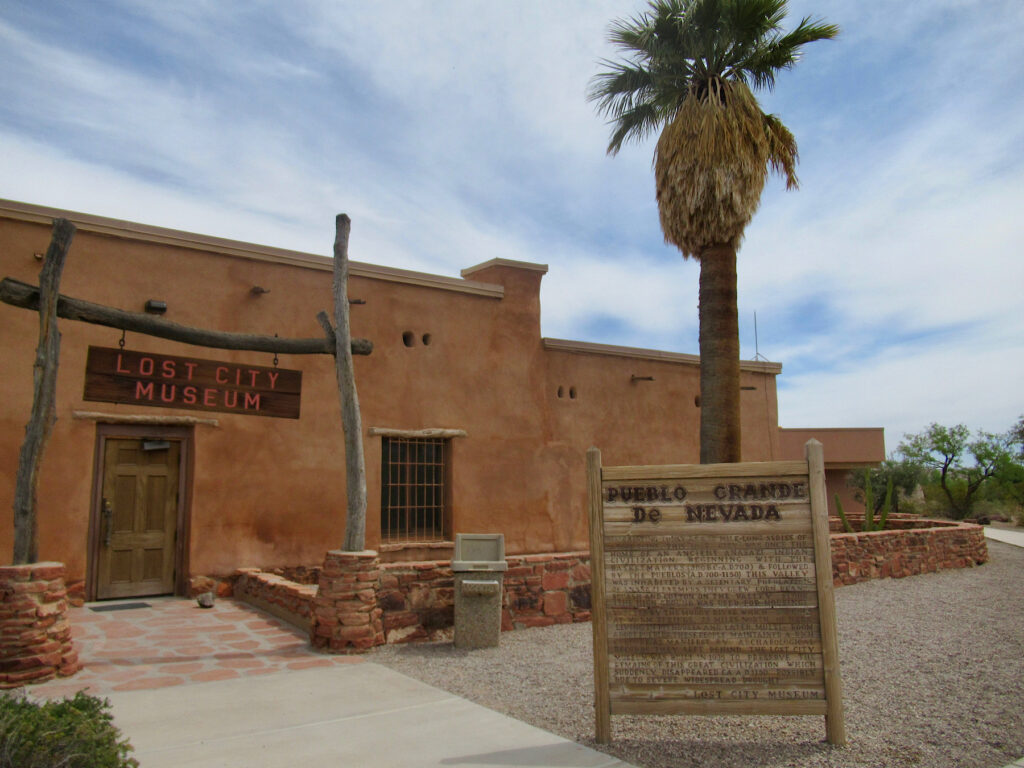 Reddish brown adobe building with sign "Lost City Museum" and lone palm tree in front.