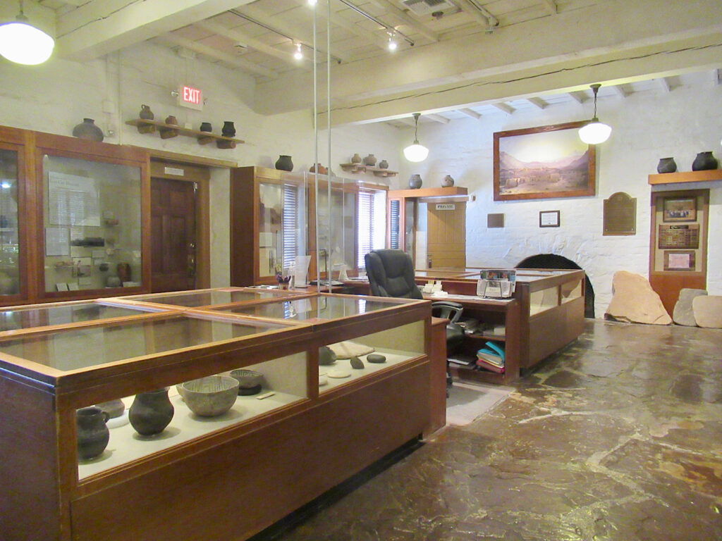 Interior of museum with white walls and white wood beam ceiling and brown stone floor. Wooden display cases full of ancient pots and old painting on wall.