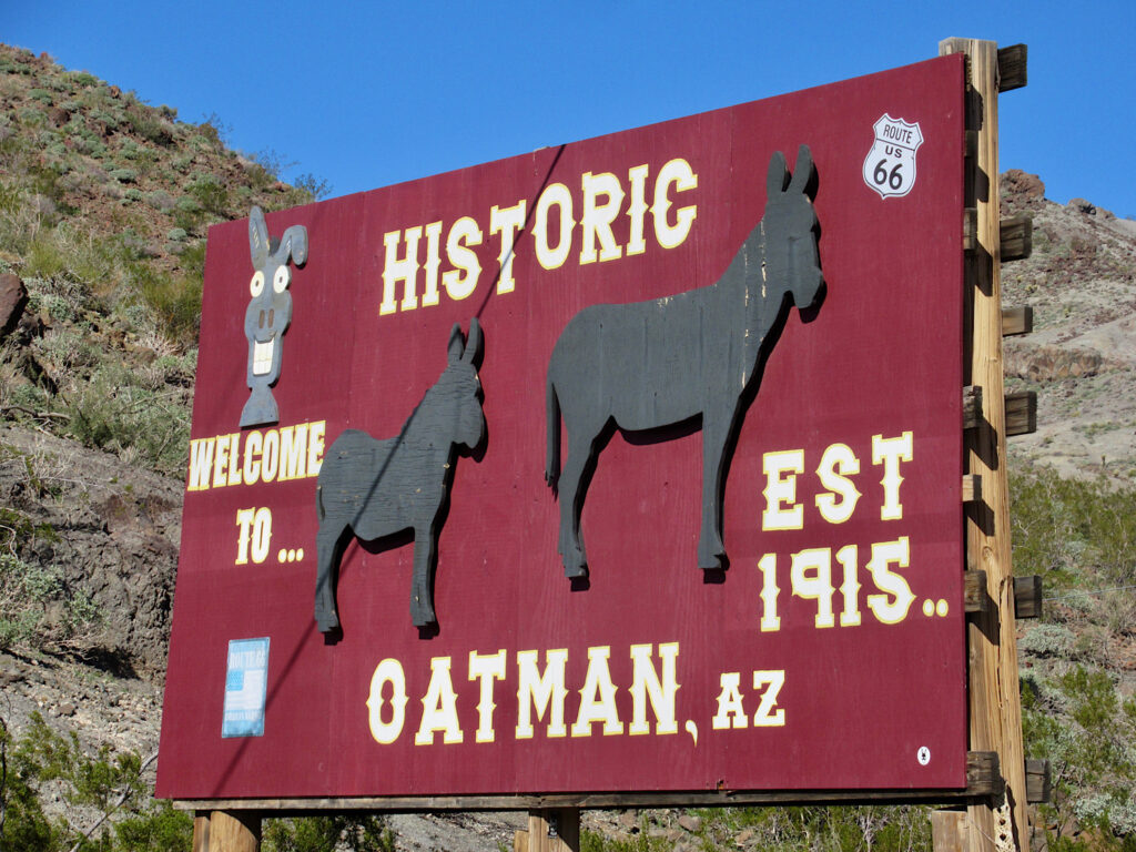Wooden, red entrance sign with images of burros reading: Welcome to Historic Oatman, AZ est 1915.