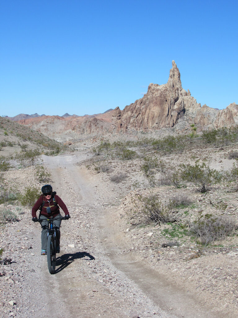 Woman on mountain bike riding on narrow gravel road with prominent pointy mountain in background under blue sky.