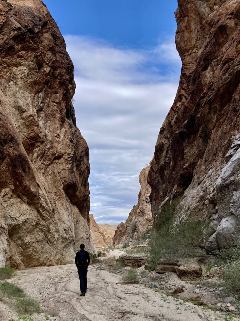 Man in black hiking up sandy wash with towering light brown canyon walls on either side below sky partially covered with clouds.