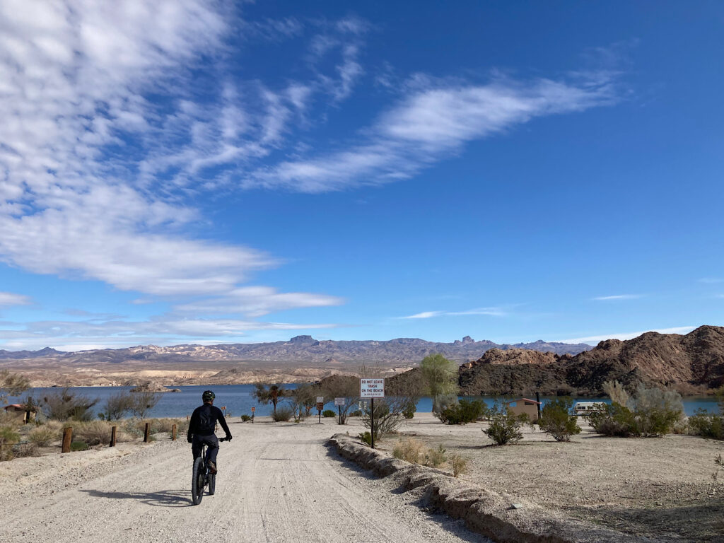 Man in black riding a mountain bike on a gravel road approaching a lake and shoreline dotted with trees.