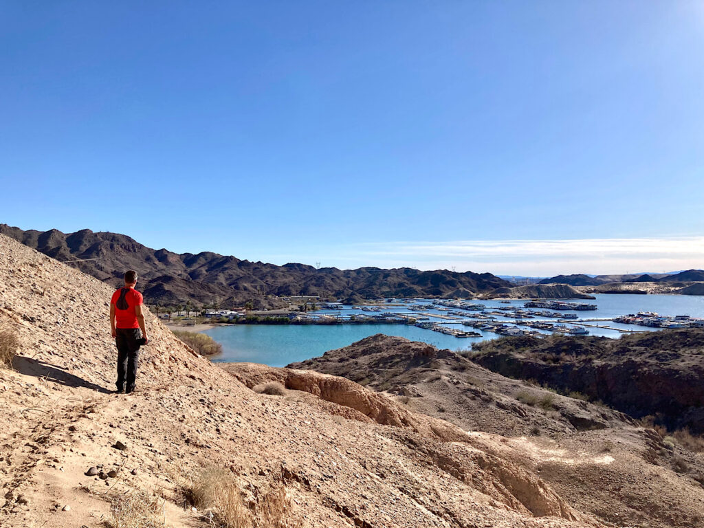 Man in red t-shirt and grey pants walking on sandy slope overlooking a marina.