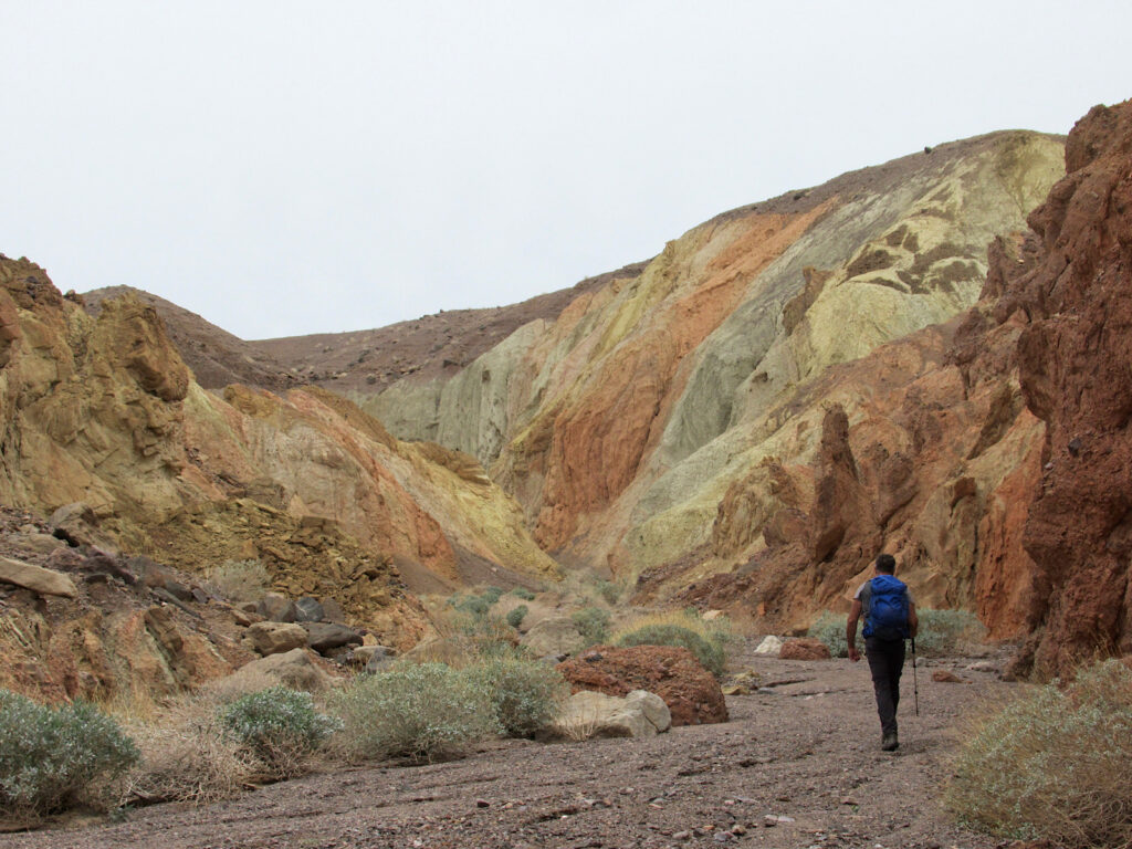 Orange, green, rusty red, brown and grey coloured slopes rising above hiking in brown-coloured wash.