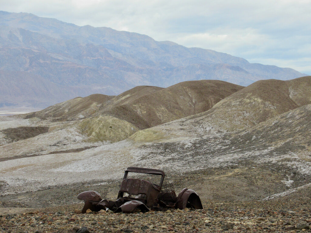Rusting frame of a Model T in foreground on rocky slope with hills and mountains in background.