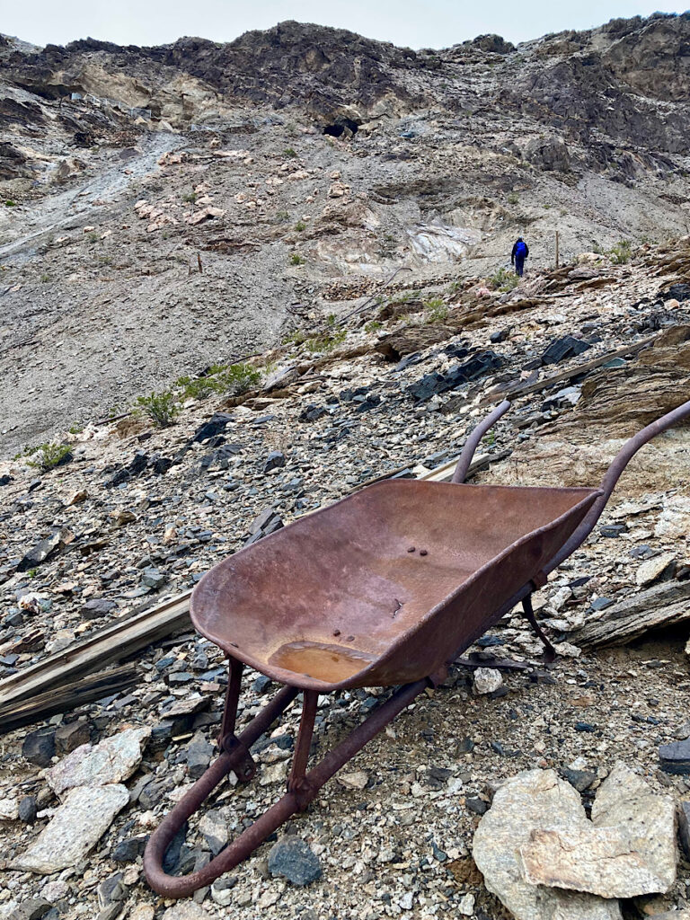 Rusty wheelbarrow frame in foreground with hiker in background heading up to ridge top.
