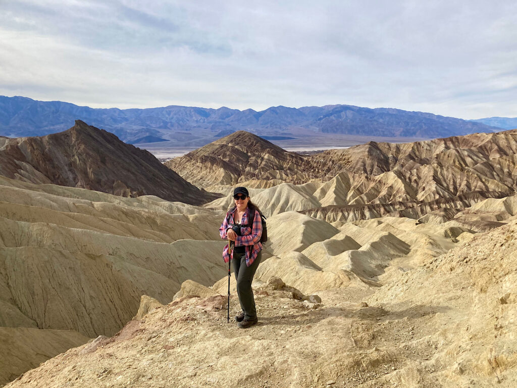 Woman in pink shirt and long black pants standing on buff coloured slop with chocolate brown hills and mountains in distance.