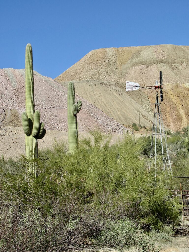 Pile of rock debris in background with a windmill and two saguaro cactus in foreground.