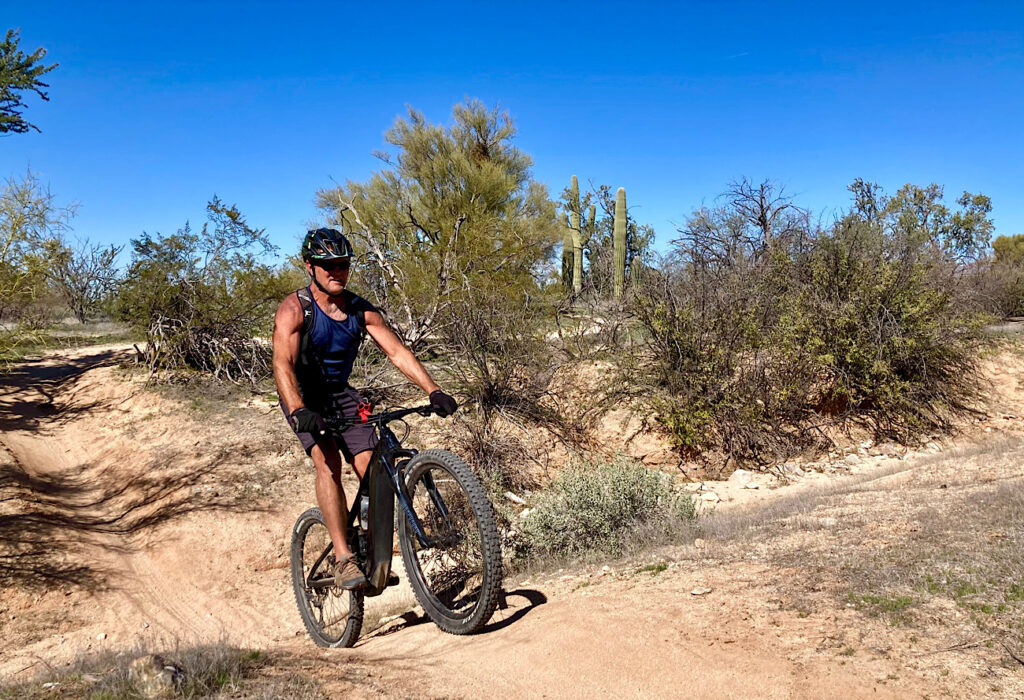 Man in blue tank top, shorts and bicycle helmet riding mountain bike on reddish dirt hill in desert.